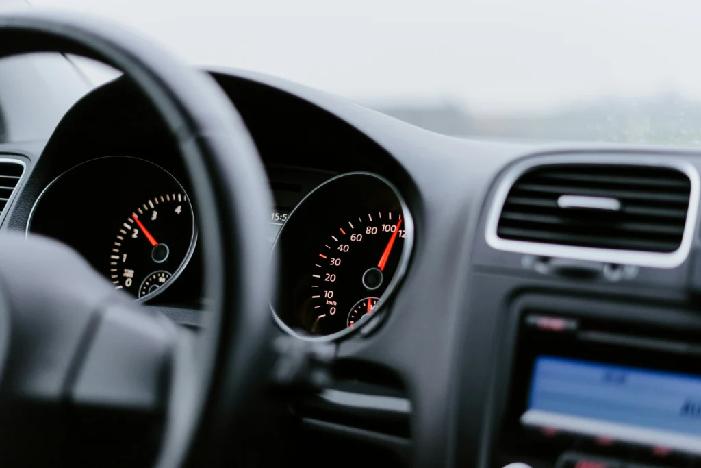 L'image montre l'intérieur d'une voiture, focalisée sur le tableau de bord et le volant. Le compteur de vitesse indique environ 100 km/h, avec un éclairage rouge sur les cadrans. L'ambiance est floue à l'extérieur, suggérant une conduite sous un ciel couvert ou pluvieux.