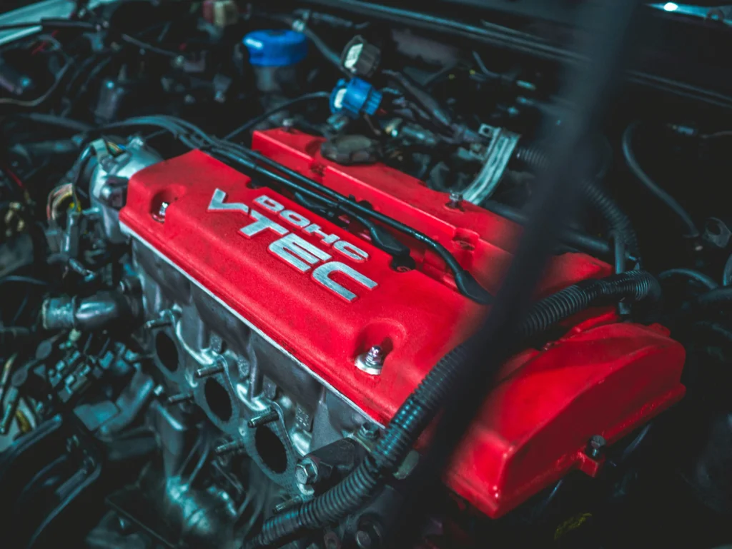 L'image montre un moteur Honda VTEC avec un cache-soupapes rouge vif portant l'inscription "DOHC VTEC". Les composants mécaniques et les câbles sont bien visibles, donnant une impression de performance et de précision. L'éclairage met en valeur les détails du moteur, soulignant son aspect puissant et sportif.